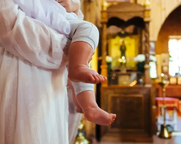 Chrzestna trzyma dziecko w kościele. W tle widać ikonostas i ławki.