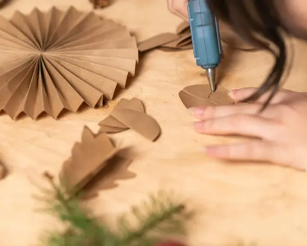 Manos creando manualidades con cartón para adultos. Se usa una pistola de silicona para pegar piezas de cartón, formando adornos.