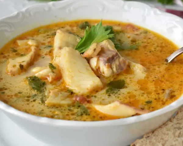 Sopa de pescado casera en un cuenco blanco con trozos de pescado, perejil y una rebanada de pan.