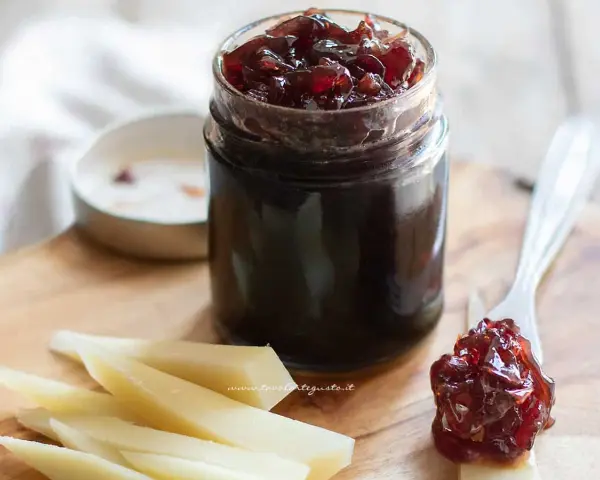 Un vasetto di marmellata di cipolle scura, con un cucchiaino che ne raccoglie una porzione, accanto a fette di formaggio su un tagliere di legno.