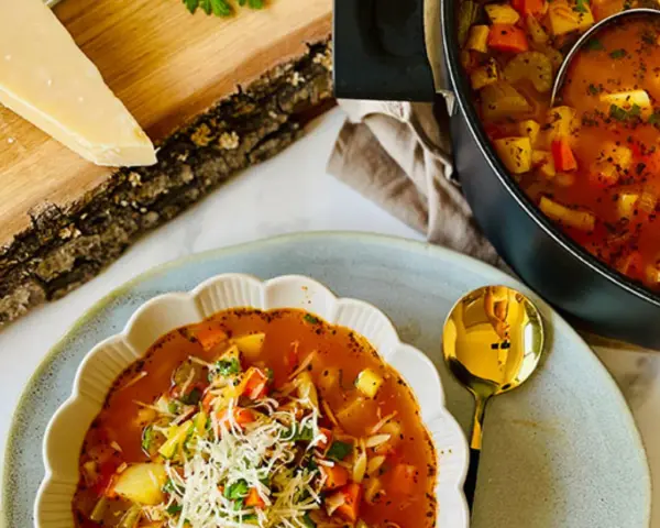 Aksamitna zupa minestrone z kawałkami warzyw i startym parmezanem, podana w ozdobnej misce. Obok garnek z zupą i kawałek sera na desce.