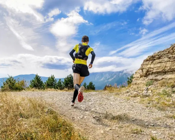 Biegacz w żółtej koszulce pokonuje kamienisty szlak. W tle góry i błękitne niebo z chmurami. To prawdziwe wyzwanie na rykowisko ultra trail.