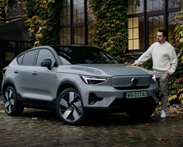 Szary Volvo C40 z czarnym dachem stoi na mokrej, brukowanej nawierzchni. Mężczyzna w białym swetrze opiera się o maskę.