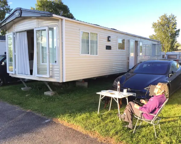 En person njuter av solen vid ett campingbord utanför en husvagn. En Tesla parkerad bredvid. Camping Timrå.