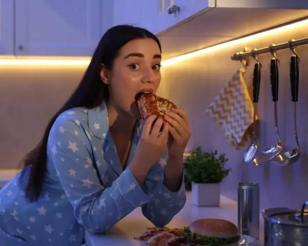 Kobieta w piżamie z gwiazdkami je kanapkę, choć czuje, że jest głodna, ale nie ma apetytu. Na blacie leży też pizza i burger.