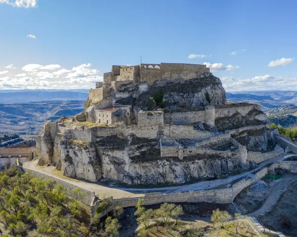 Castillo de Morella sobre un peñón rocoso, con murallas y edificios de piedra bajo un cielo azul con nubes.