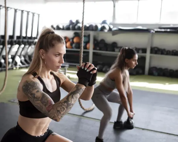 Dwie kobiety wykonują przysiady z kettlebellami. Intensywny trening funkcjonalny w siłowni.