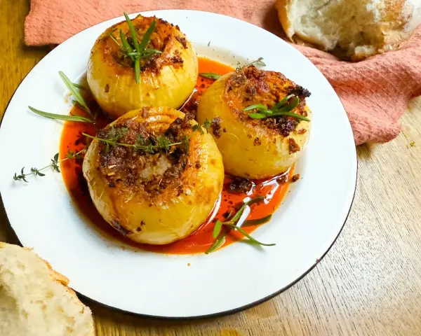 Tres cebollas al horno, rellenas y doradas, nadan en una salsa roja vibrante. Hierbas frescas coronan cada una.