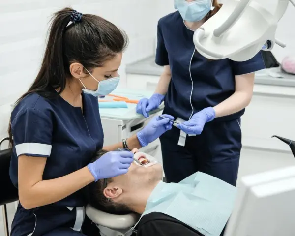 Dwie dentystki w maseczkach i rękawiczkach wykonują zabieg stomatologiczny u pacjenta.
