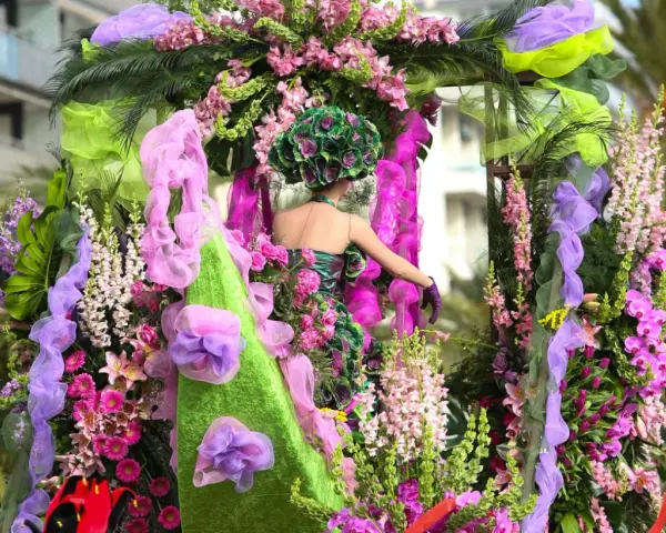 Una figura con un vestido floral y un tocado verde, rodeada de exuberantes flores rosas y moradas, participa en el **carnaval de Nice**.