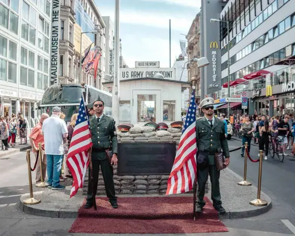 Zwei Männer in Uniformen mit US-Flaggen stehen am Checkpoint Charlie in Berlin. Touristen und ein McDonald's im Hintergrund.