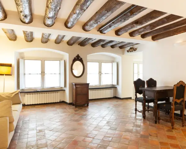 Stary salon z belkami stropowymi i podłogą z terakoty. Kanapa, stół z krzesłami i komoda tworzą przytulną atmosferę.