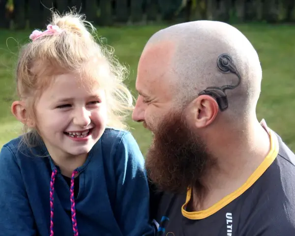 Uśmiechnięta dziewczynka i mężczyzna z tatuażem implantu ślimakowego na głowie. Rozmowa o implancie ślimakowym za i przeciw.