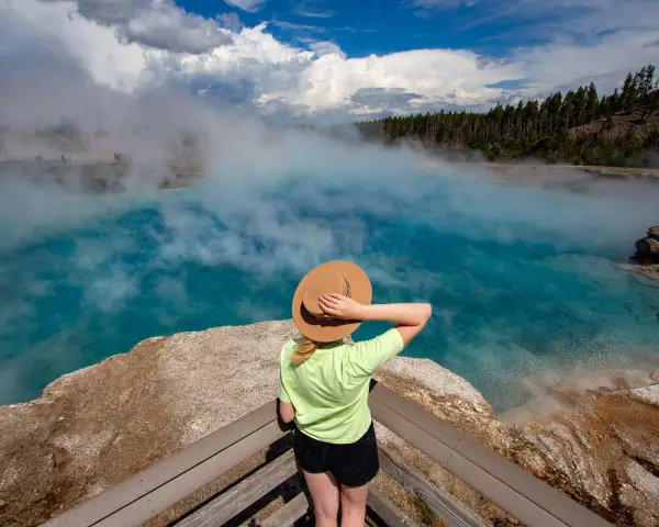 Kobieta w kapeluszu podziwia turkusowe gorące źródło w Yellowstone. Gdzie jest Yellowstone? Tu, wśród parujących wód i lasów.
