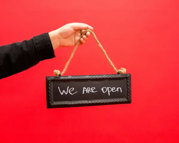 Mano con chaqueta negra sostiene un cartel que dice "We are open", indicando la apertura de un negocio, similar a los tipos de licencia de actividad.