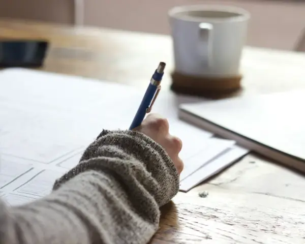 Ręka w swetrze pisze długopisem po papierze. Obok kubek i notatnik. Co to za część mowy?