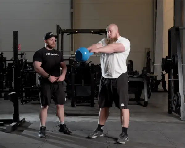 Man med skägg utför kettlebell swing varje dag i gym. Bredvid står en tränare.