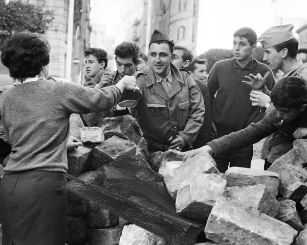 Un grupo de jóvenes y soldados interactúan tras una barricada de piedras en Argelia. Un hombre con uniforme militar sirve bebida.