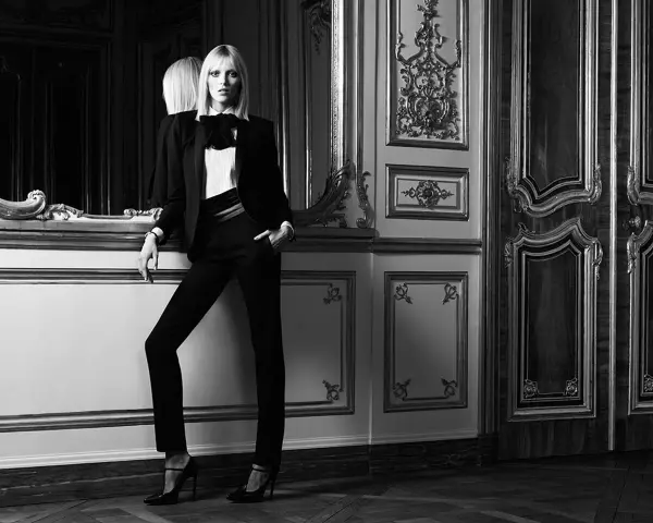Donna elegante in un impeccabile outfit smoking, con giacca nera, camicia bianca e papillon, posa con sicurezza in un lussuoso salone.