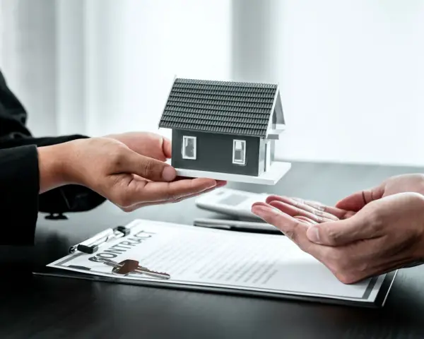 Manos entregan maqueta de casa sobre contrato y llaves. Posible exención IRPF venta vivienda heredada.