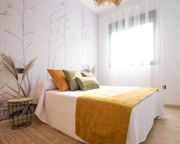 Habitación con papel tapiz de ramas y gotele moderno. Cama con cojines verdes y naranjas, y manta mostaza.