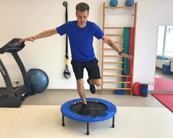 Mężczyzna wykonuje ćwiczenia po rekonstrukcji ACL, balansując na małej trampolinie. W tle bieżnia i drabinka gimnastyczna.