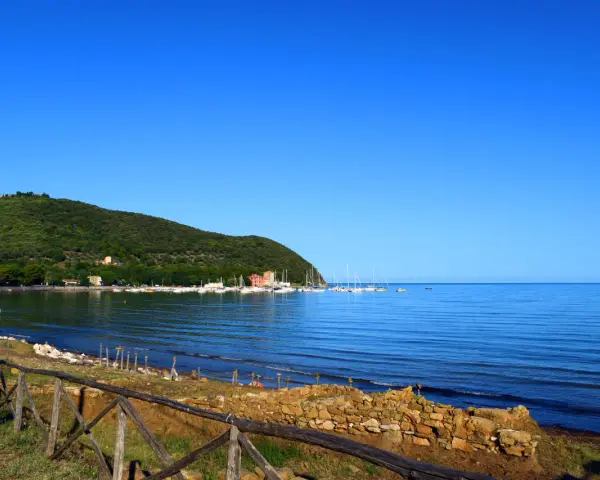 A tranquil bay in Tuscany with sailboats docked, a green hillside, and a rustic wooden fence in the foreground.