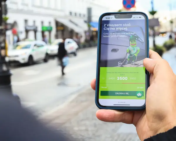Dłoń trzyma telefon z aplikacją Vivus, gdzie można złożyć wniosek o pożyczkę. W tle miejski krajobraz. Vivus windykacja pomaga spełniać marzenia.