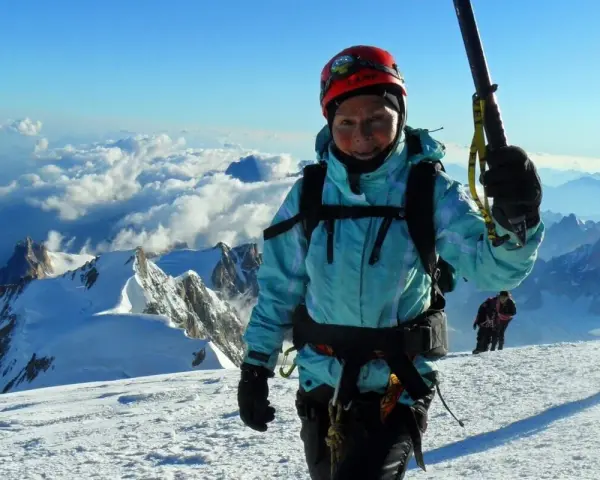 Barbara Prymakowska, mimo swojego wieku, zdobywa szczyty. Uśmiechnięta alpinistka w kasku i turkusowej kurtce na tle gór i chmur.