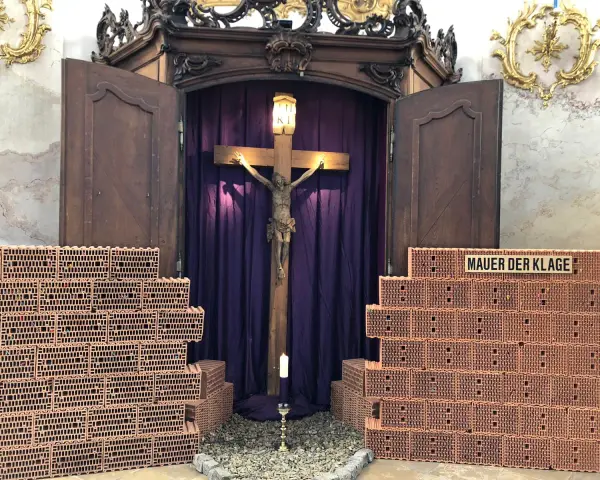 Kreuzweg Stationen: Jesus am Kreuz, umgeben von Ziegelmauern, die die "Mauer der Klage" symbolisieren. Eine Kerze brennt vor dem Kruzifix.