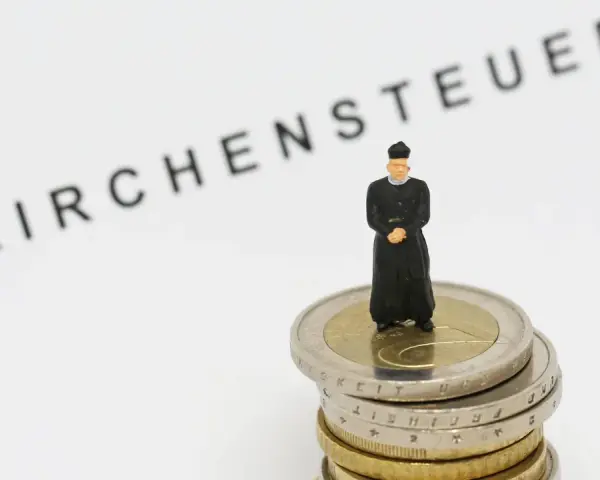 Miniatur-Priester steht auf Münzstapel. Der Schriftzug "Kirchensteuer" im Hintergrund wirft die Frage auf, warum man kirchensteuer zahlt.