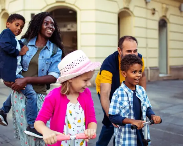 Glückliche Familie unterwegs: Mutter trägt Kind, Vater schiebt Roller. Ein Spagat zwischen Kind und neuem Partner gelingt hier spielerisch.