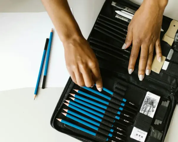 Un étui ouvert révèle une sélection de crayons de différents types pour dessiner, avec des mains choisissant un crayon bleu.