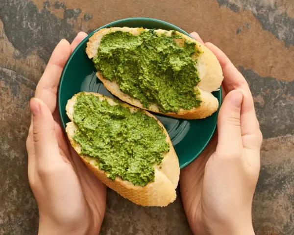 Dwie kromki bagietki z aromatycznym pesto z pietruszki na zielonym talerzyku, trzymanym w dłoniach.