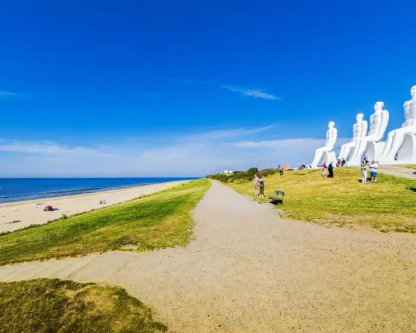 De vita sandstränderna i Danmark möter en grönskande sluttning med gigantiska sittande figurer. En grusväg leder mot havet under en klarblå himmel.