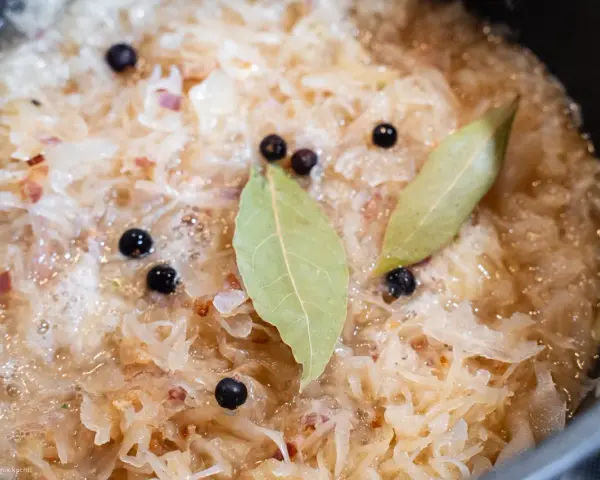 Sauerkraut köchelt mit Lorbeerblättern und Wacholderbeeren, um den Geschmack zu verfeinern. Kleine rote Zwiebelstücke sind ebenfalls sichtbar.