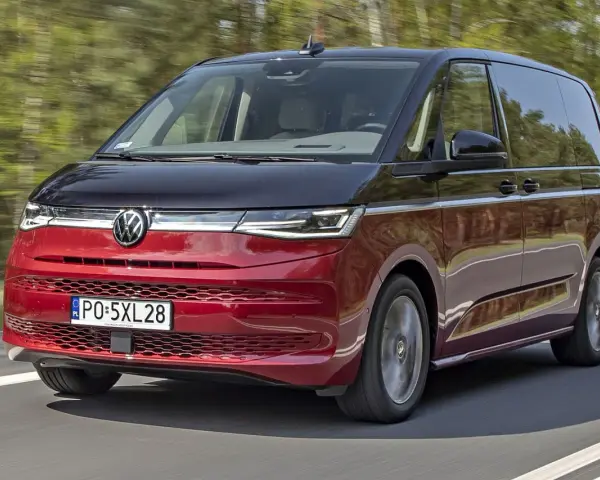 Nowy Volkswagen van w kolorze czerwono-czarnym pędzi po drodze, otoczony zielenią drzew.