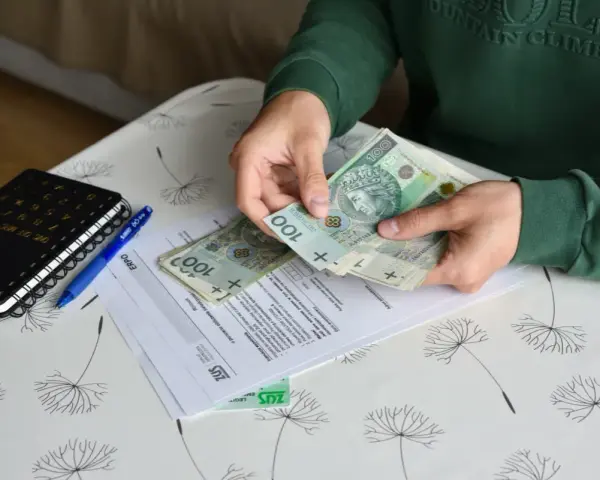 Ręce liczą banknoty 100 zł nad dokumentami ZUS. Obok leży kalkulator i długopis. To może być pomocne przy ustalaniu, ile wynosi świadczenie przedemerytalne na rękę.
