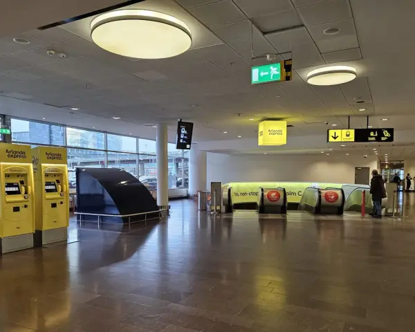 Żółte automaty biletowe Arlanda Express na lotnisku w Sztokholmie. W tle ruchome schody i znaki kierujące.