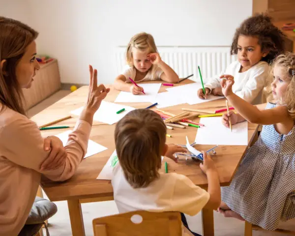 Bambini disegnano e giocano con un aereo, guidati da un'insegnante. Un esempio di come le scienze della formazione sbocchi in attività creative e formative.