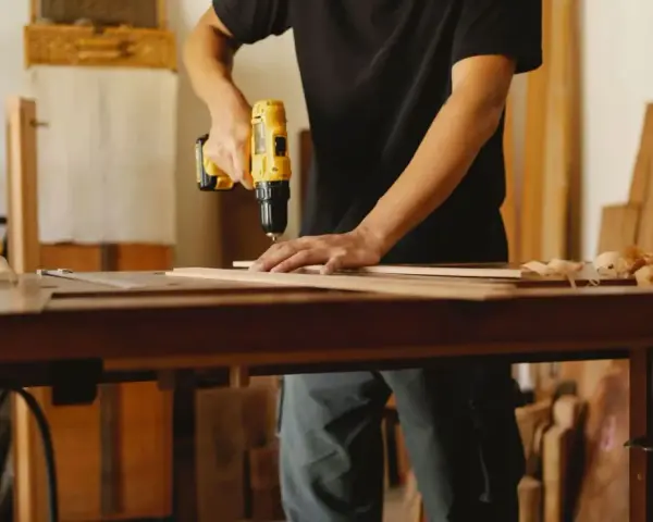 Mann bohrt Holzbretter, um seine Werkstatt einzurichten. Hobelspäne liegen auf dem Tisch.