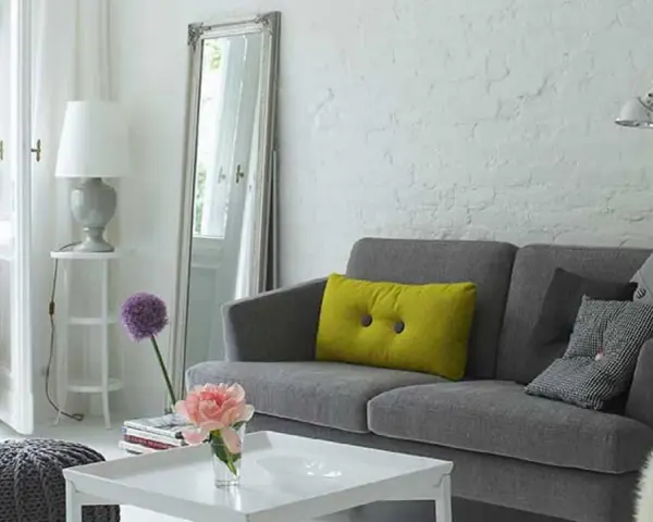 Sofá gris con cojines, espejo alto y lámpara. Trucos para decorar espacios pequeños con estilo y amplitud.