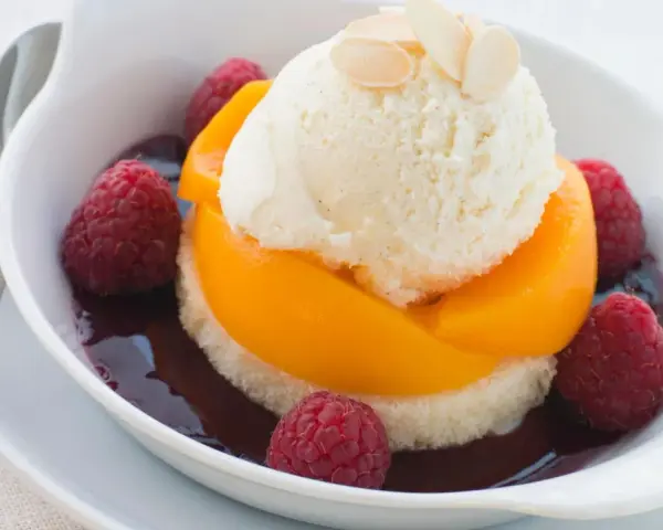 Ein köstlicher Pfirsich Melba: Vanilleeis auf Pfirsichspalten, umgeben von Himbeeren und dunkler Sauce, garniert mit Mandelblättchen.