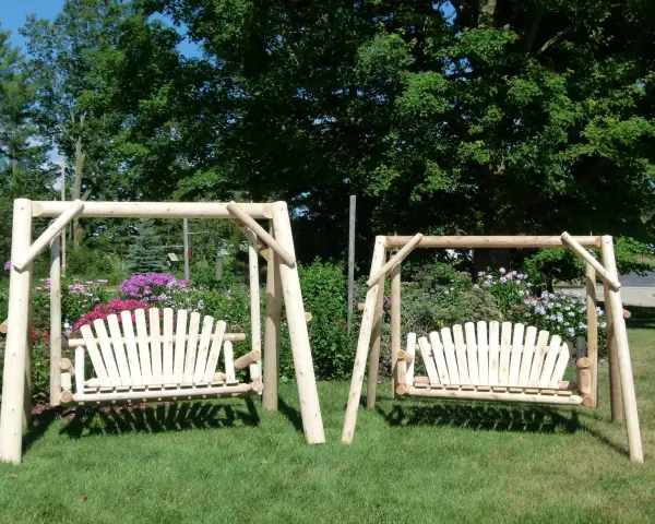 Two wooden porch swings stand on a grassy lawn, surrounded by colorful flowers and lush trees. This image offers inspiration for how to hang a porch swing.