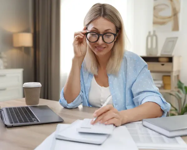 Femme aux lunettes calculant, laptop et café à proximité. Elle semble gérer des finances, peut-être une réduction de capital non motivée par des pertes.