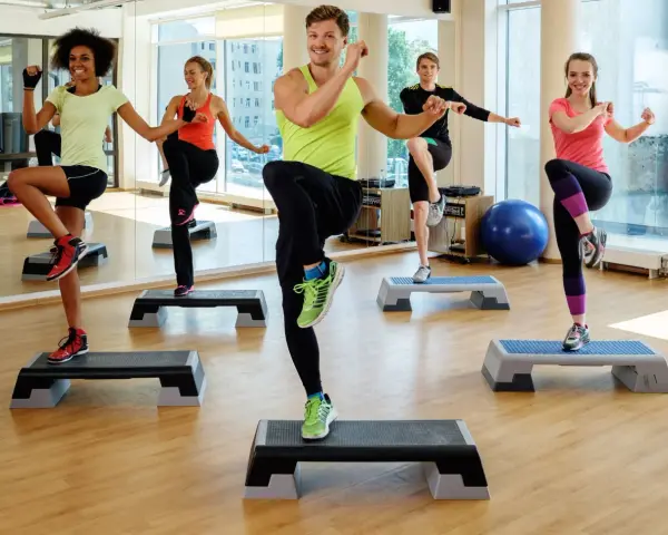 En grupp människor utför step ups i ett gym. De har roligt medan de tränar.