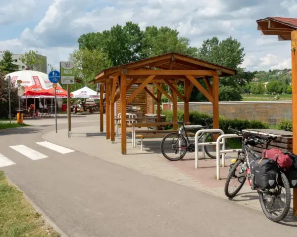 Miejsce odpoczynku przy ścieżce rowerowej. Dwa rowery, w tym jeden z sakwami, czekają na dalszą podróż. W tle zadaszenie i stoliki, idealne na przerwę w trasie.