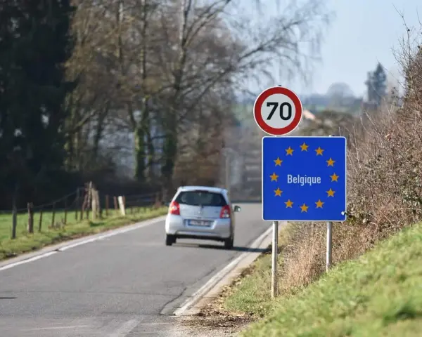 Znak graniczny Belgii, gdzie potrzebny paszport, na tle drogi i jadącego samochodu.