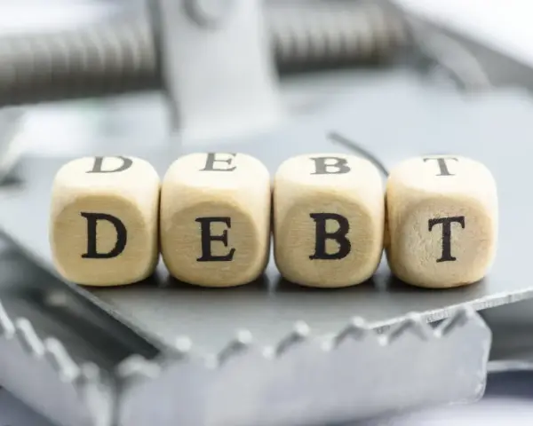 Cztery drewniane kostki z literami D, E, B, T tworzą słowo "DEBT" na pułapce. To symbol finansowych zobowiązań, które mogą prowadzić do trudnej sytuacji, jak datio in solutum.