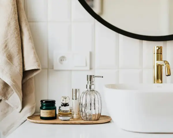 Sur un plateau en bois, des flacons de parfum et un distributeur de savon argenté. Une serviette beige est suspendue à côté d'un lavabo blanc et d'un robinet doré. La colle carrelage flex, c'est quoi ?